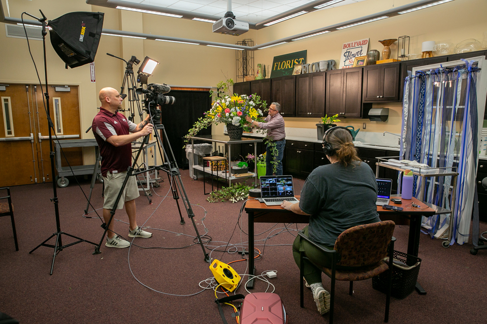 A person arranges flowers in the center of a studio while another person records with a video camera and another works the soundboard.