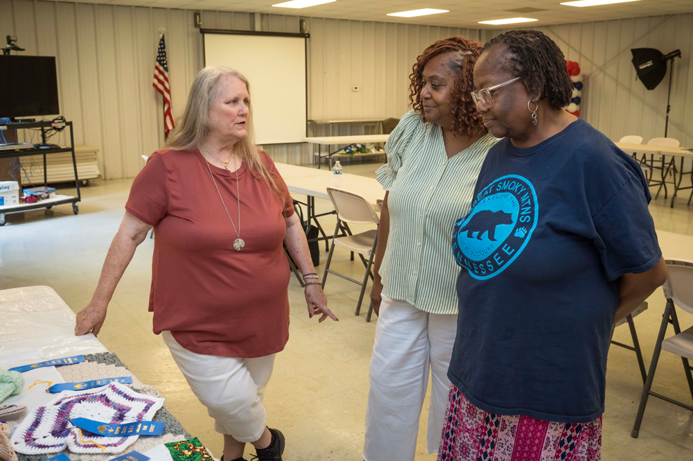 A person talks to two others who examine a table with homemade crafts and award ribbons.