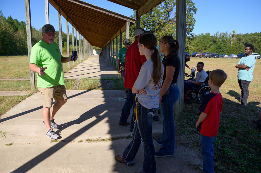 A man in a green shirt gestures toward a group of teens at a shooting range.