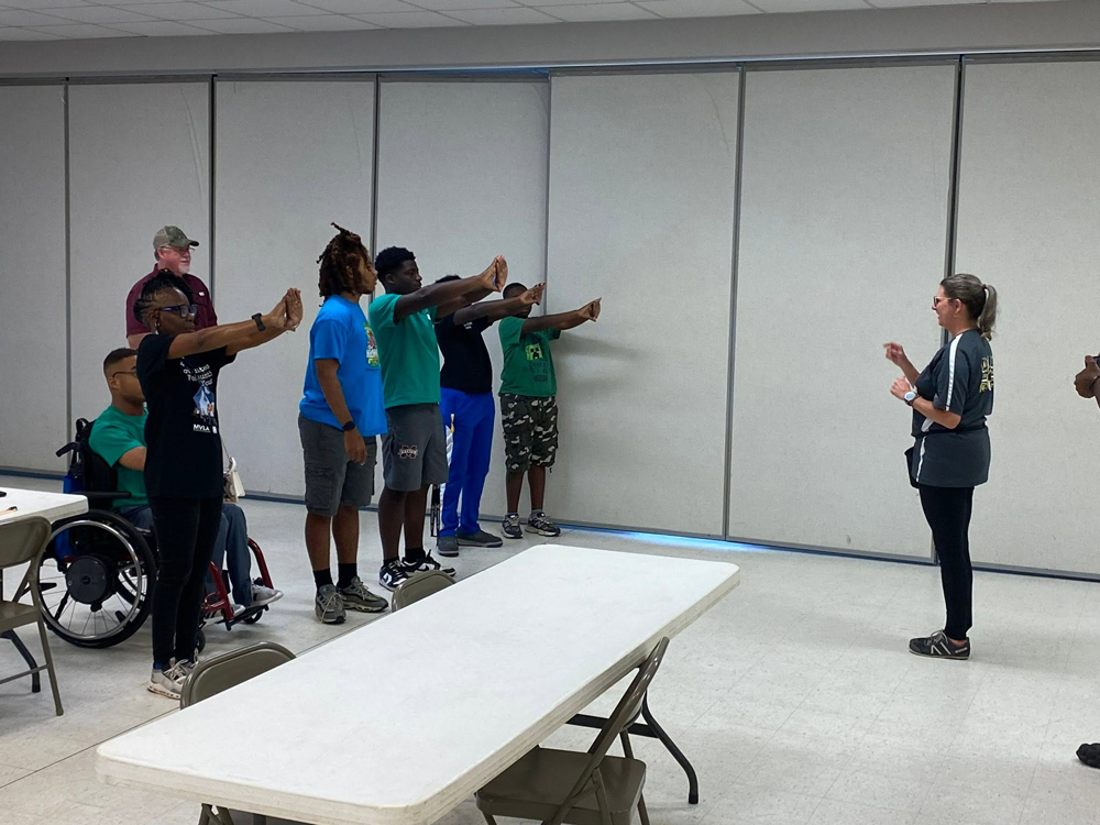 One adult points at six teens doing stretching exercises as another adult looks on.