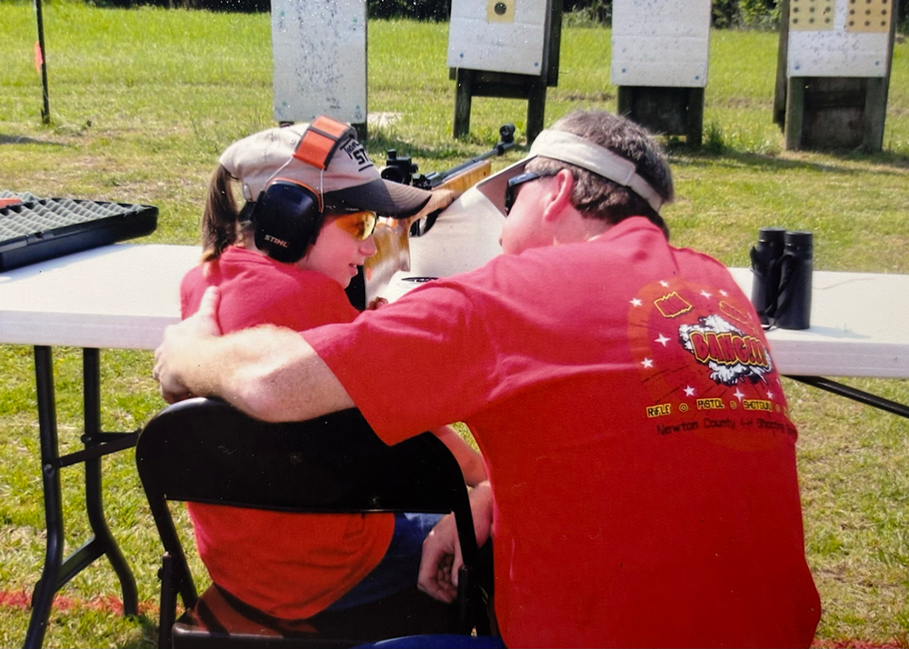 An adult gives a side hug to a smiling child at a shooting range.