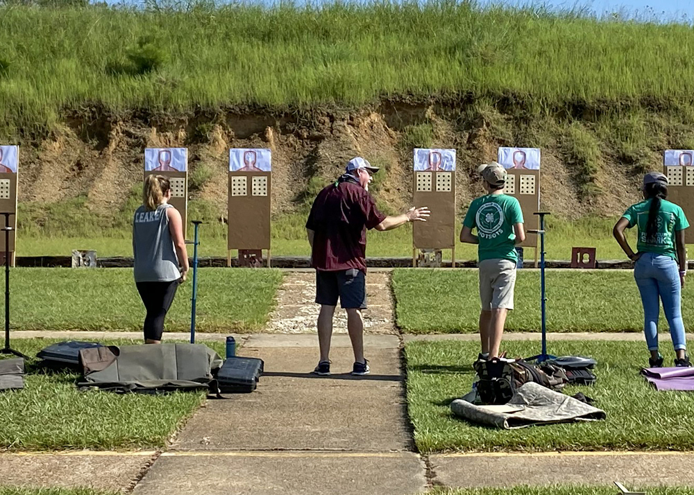 A group of three teens standing outside at a shooting range and looking at a gesturing man.