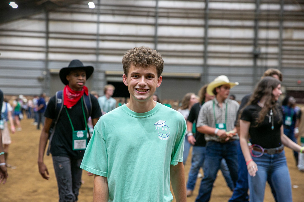 : A teen smiling and dancing as other teens line dance behind him in a horse barn. 