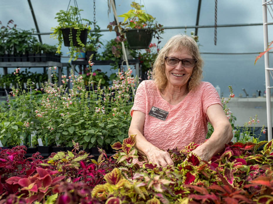 A person smiling in a greenhouse surrounded by colorful foliage.