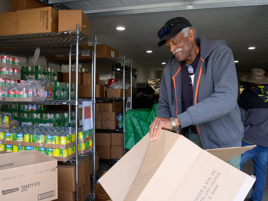 A man flattens an empty cardboard box beside metal shelving units stocked with canned goods and boxed items in a food pantry.