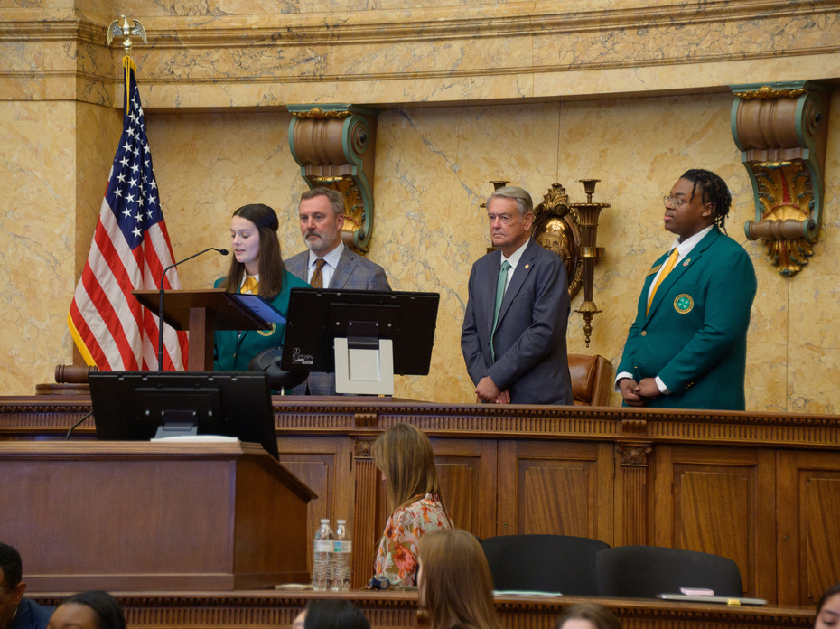 A teenager speaks at a podium as two adults and another teen look on.