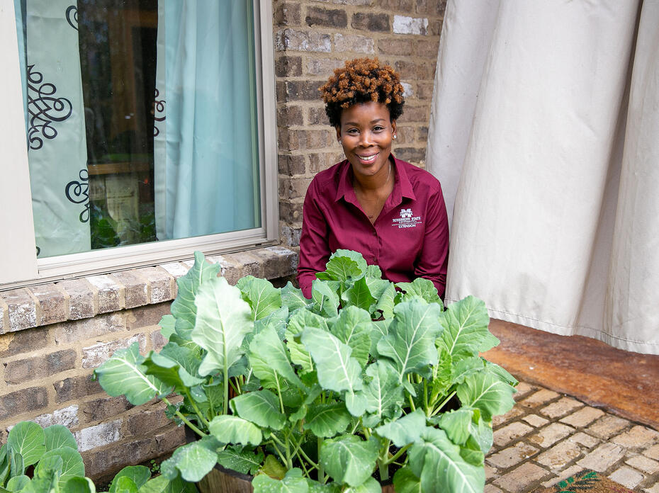 A woman kneels behind a planter on a brick patio.