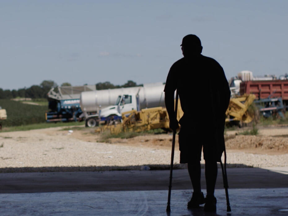 A man stands on crutches in silhouette against a background of farm equipment.