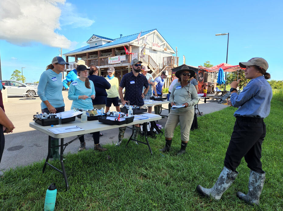 A woman talks to a group gathered at a table outdoors.