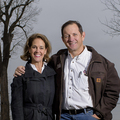 A man stands next to a woman with his arm around her shoulders while they both smile at the camera; a lake rests in the background.
