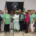 A group of 14 men and women stand on either side of a woman wearing a bright green hat and holding up an award.