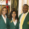 Two young women and one young man wearing green blazers stand on a marble floor.
