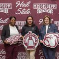 Three women standing in front of a maroon and white Mississippi State University banner.