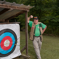 A smiling man wearing a vest and green shirt, standing beside an archery target with three arrows near the bullseye.