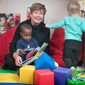 A woman reads to one child on her lap while another walks nearby.