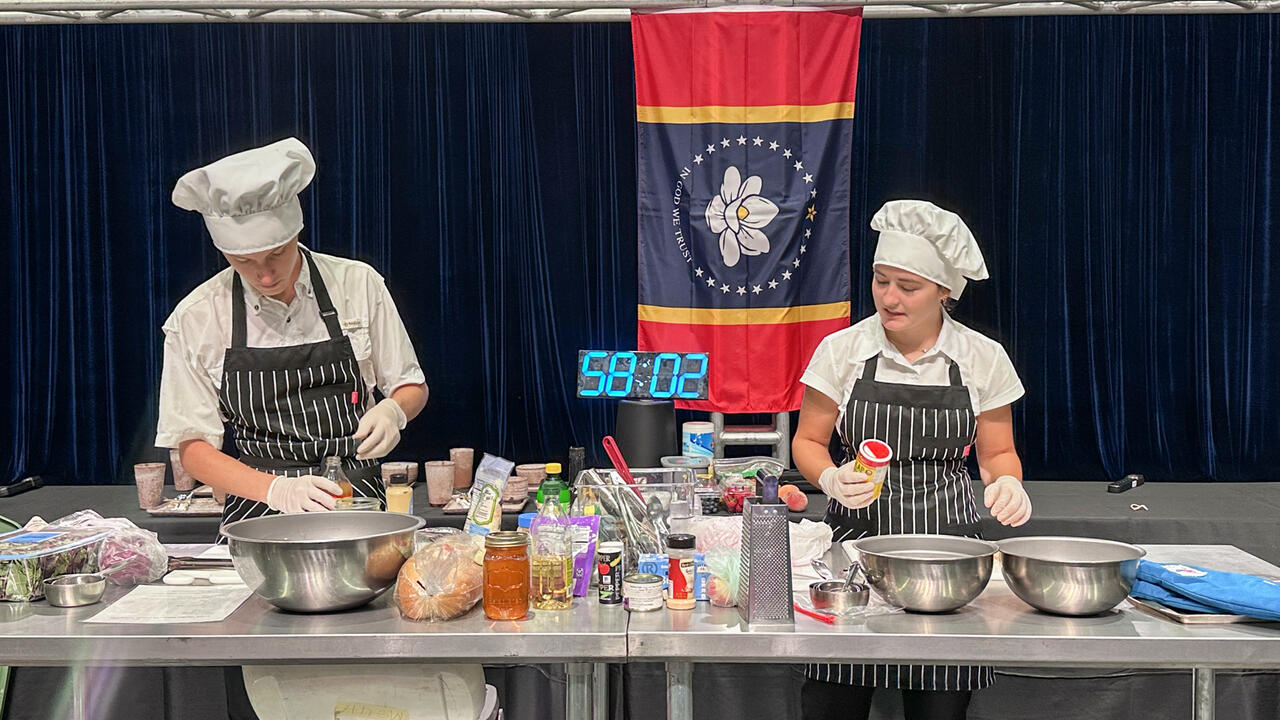 Two people prepare food during a cooking competition with a large timer in the background.
