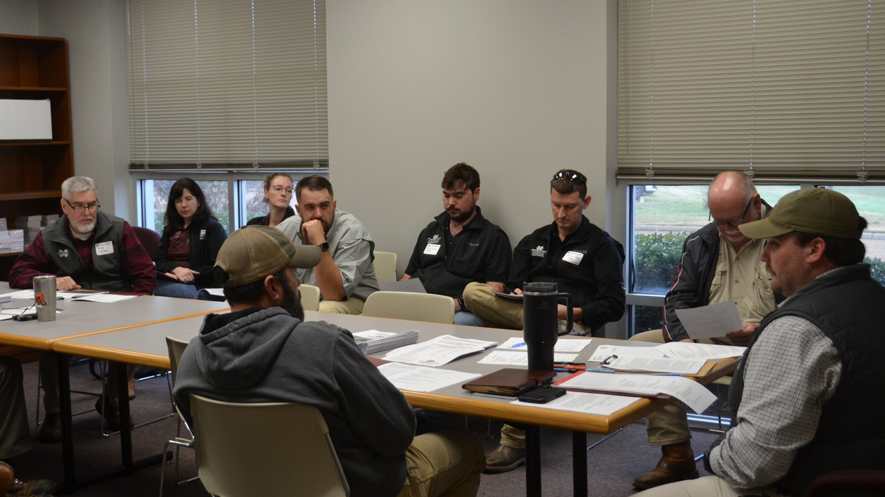 A man sits at the end of a conference room table with others gathered around.