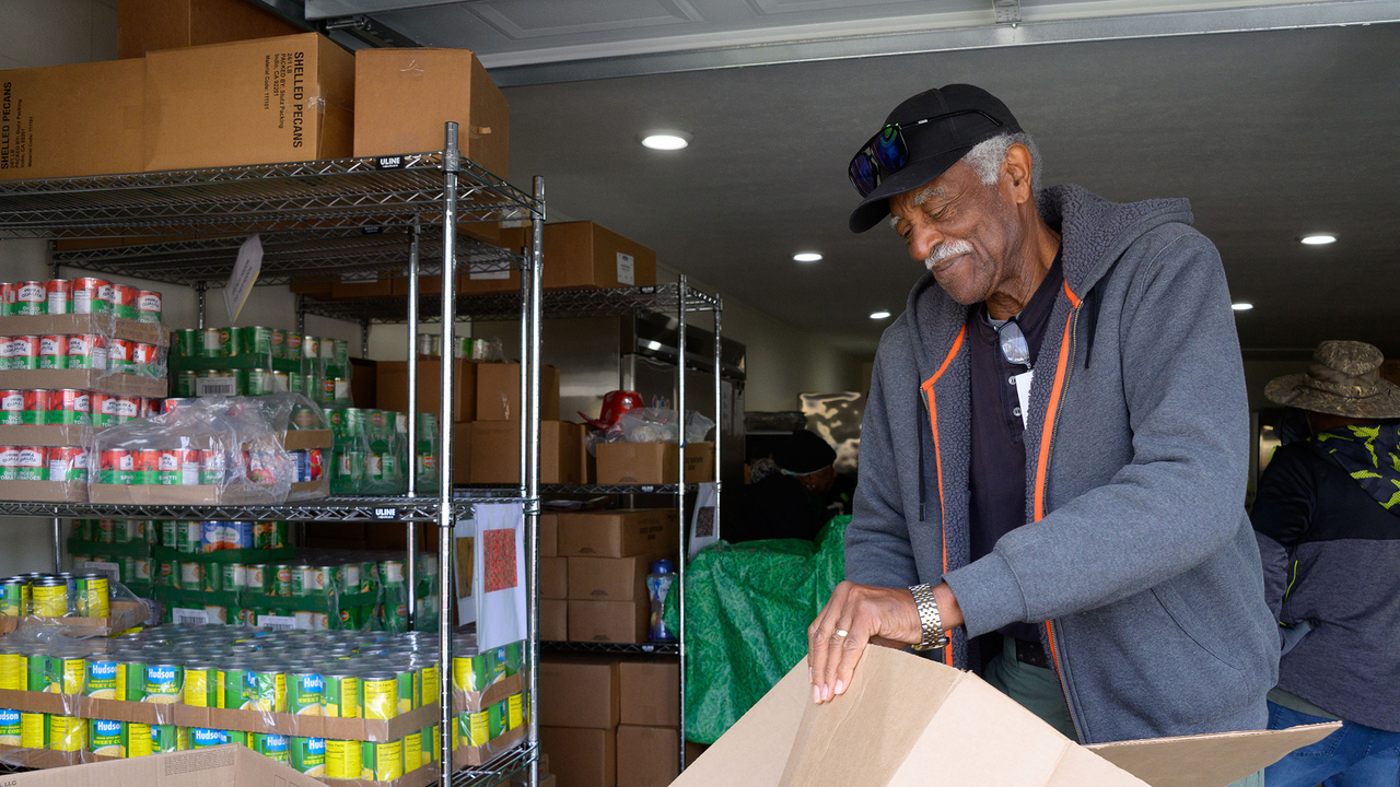 A man flattens an empty cardboard box beside metal shelving units stocked with canned goods and boxed items in a food pantry.