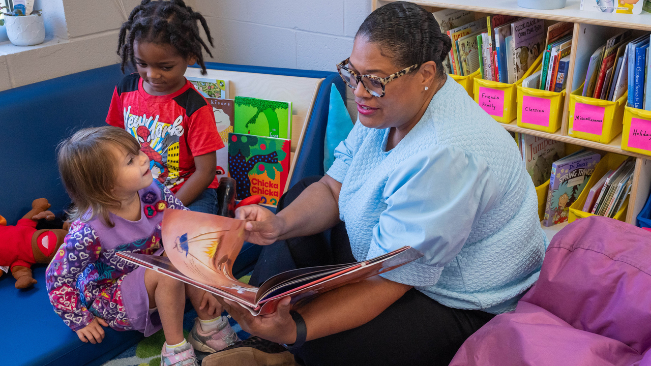 An adult reads from a picture book to two young children seated in a classroom reading corner.