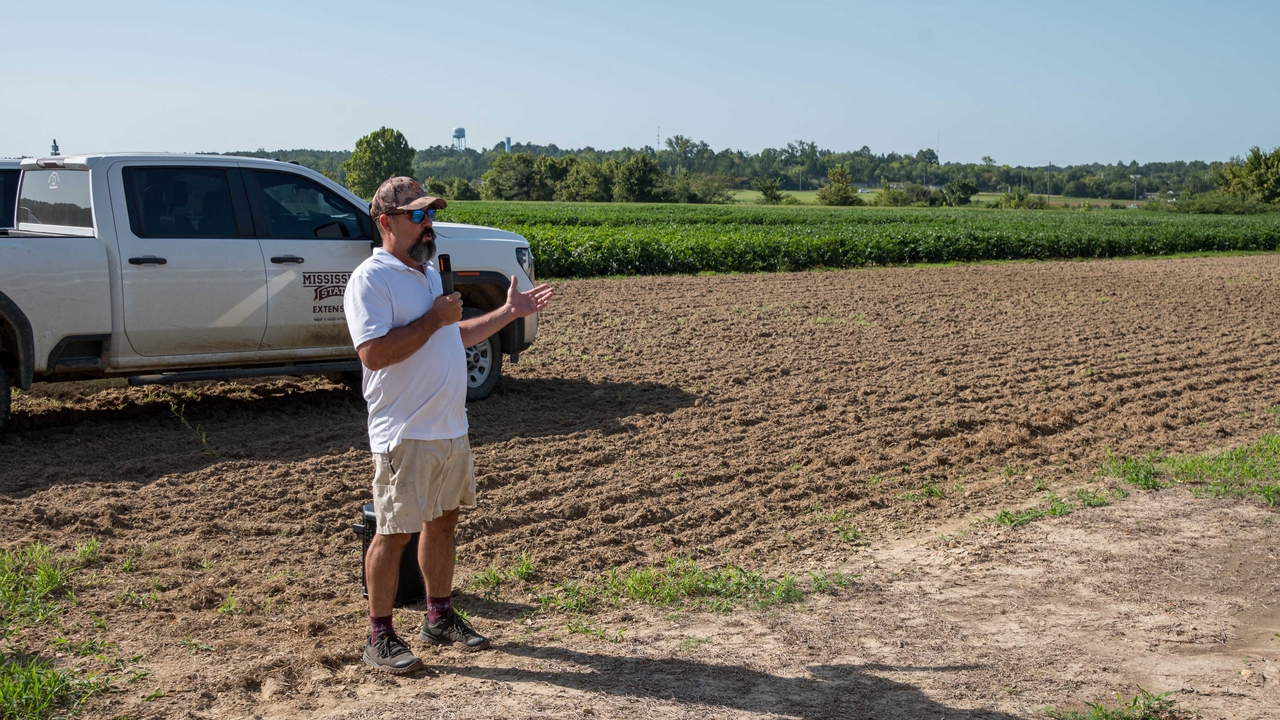 A speaker stands in front of research plots with a Mississippi State University Extension pickup truck in the background.