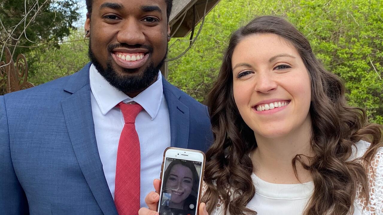 A smiling couple holds a smart phone showing a woman on a video call.