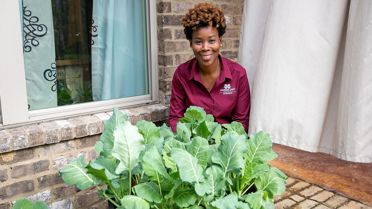 A woman kneels behind a planter on a brick patio.