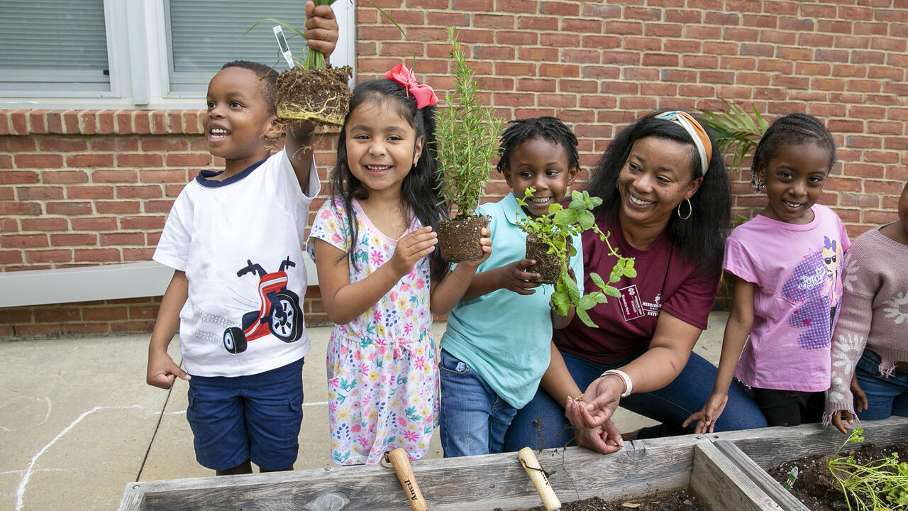 Four students stand in a row with their teacher and hold up the herbs they were about to plant in the raised bed.