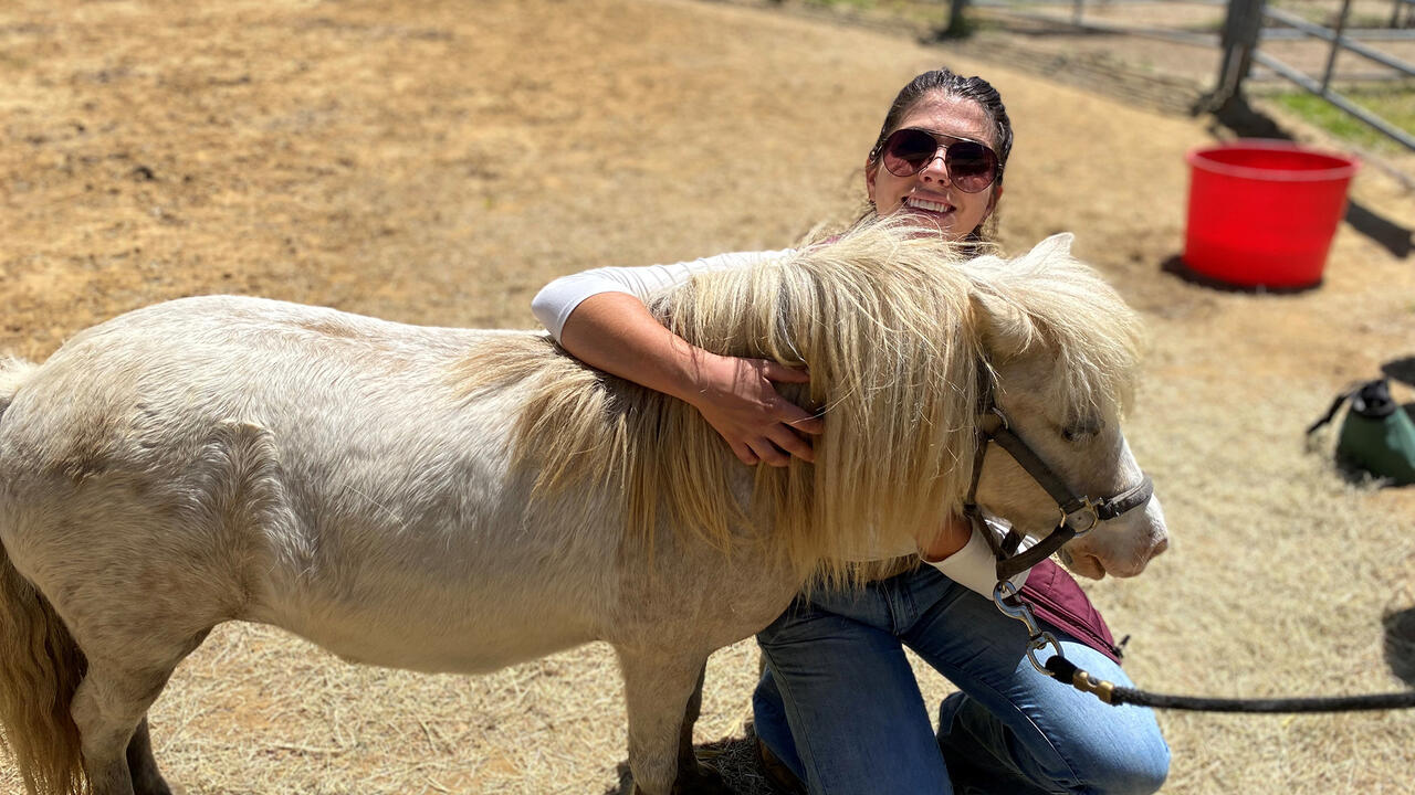 A woman kneels beside a small horse that she is hugging.
