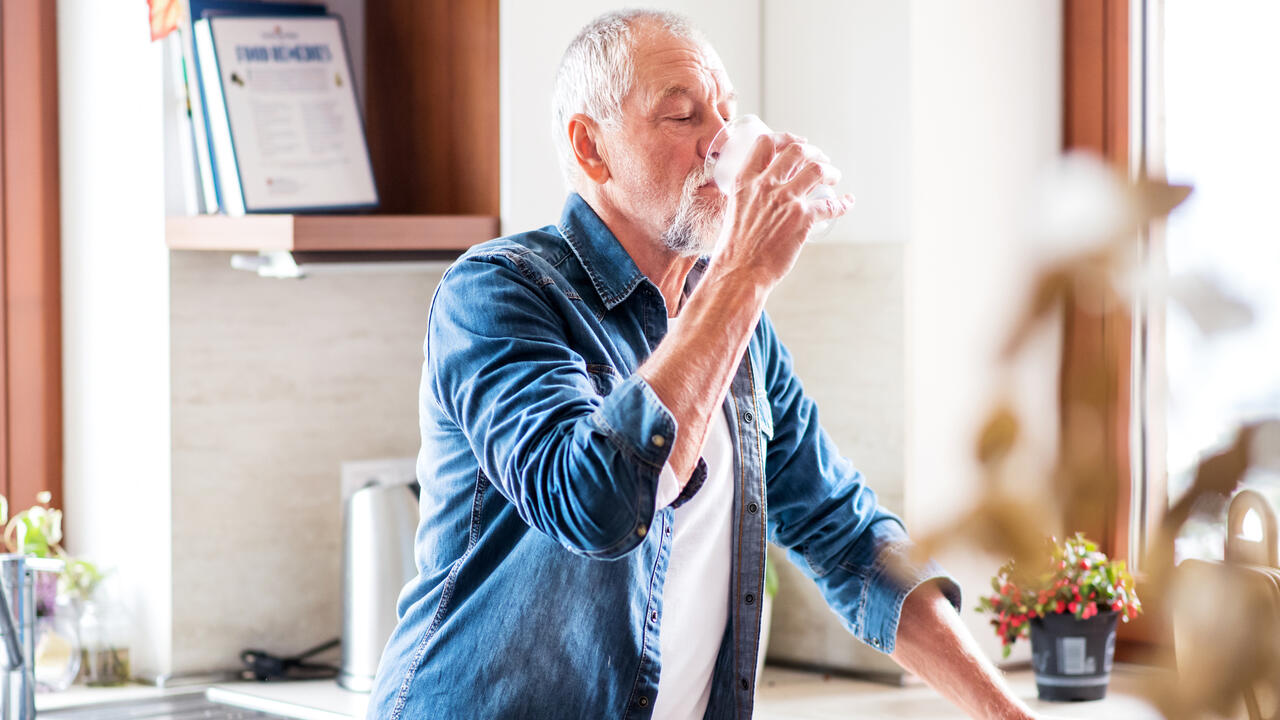 An older man drinks water in the kitchen.