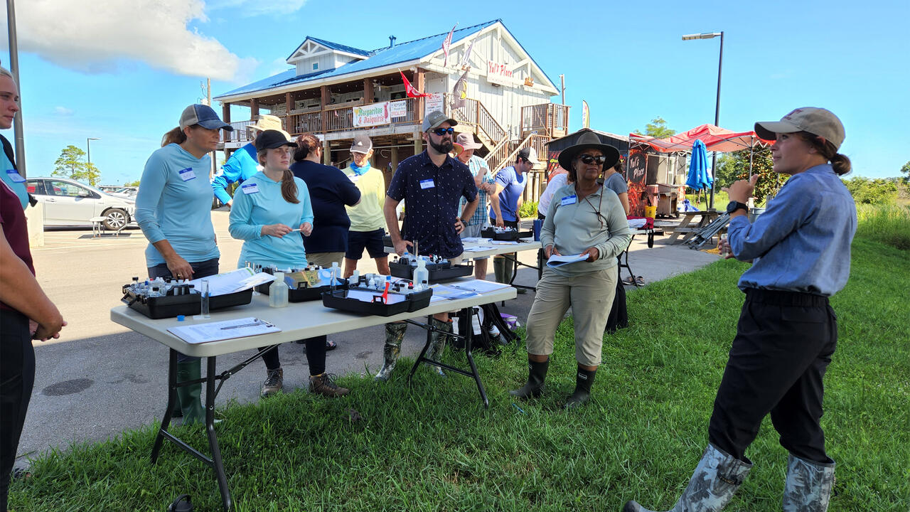 A woman talks to a group gathered at a table outdoors.