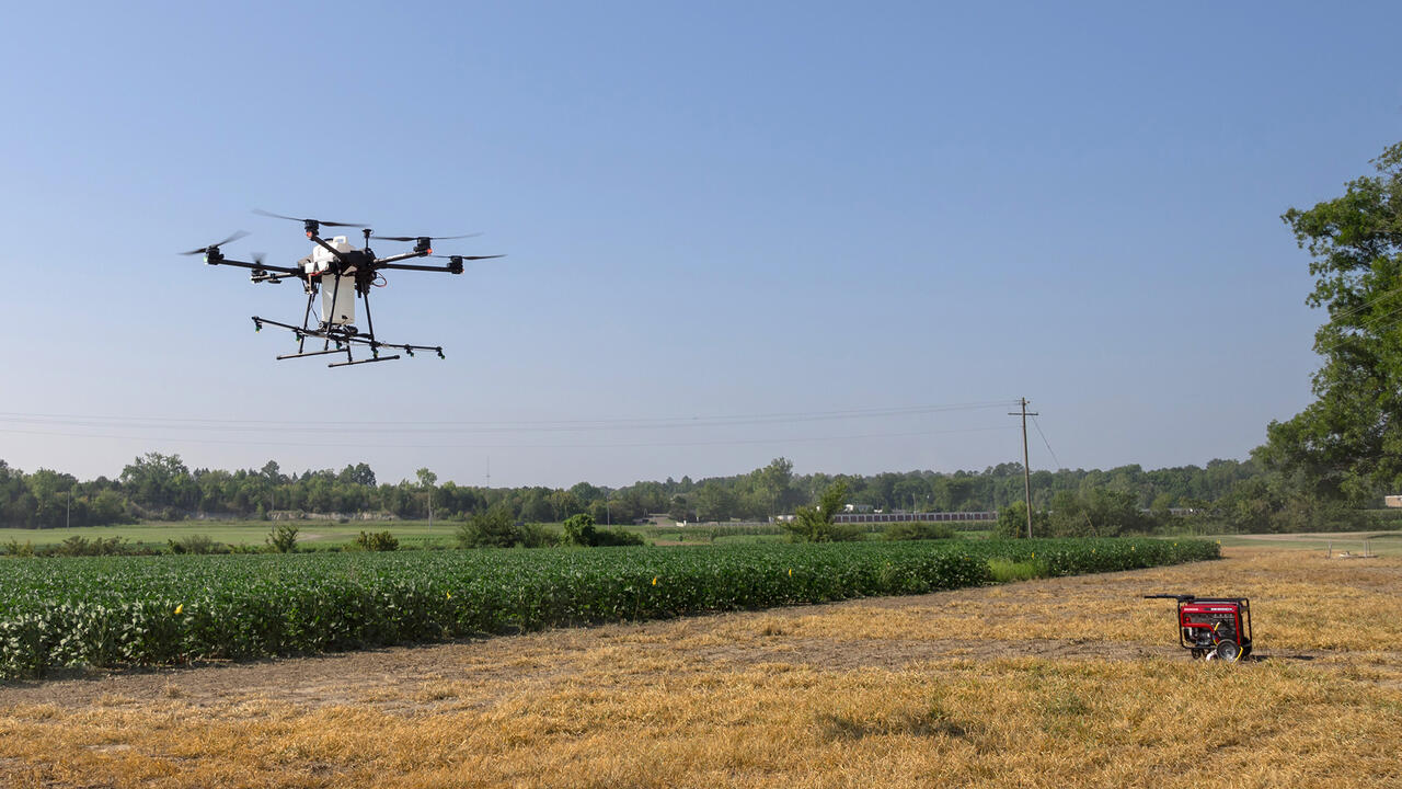 A large drone hovers over cropland.