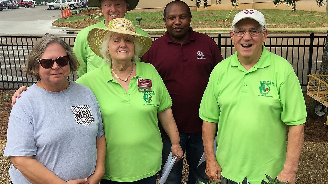 A group of five adults stand in front of some plants.