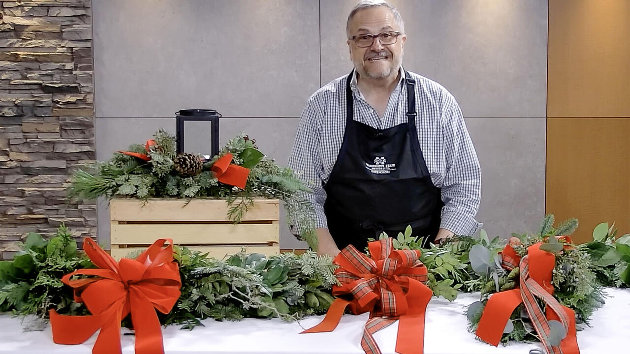 A man stands in a studio with a completed velvet bow, garland, door swag, and table centerpiece.