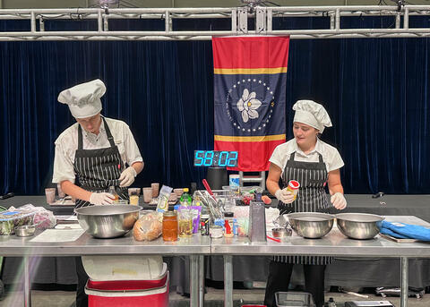 Two people prepare food during a cooking competition with a large timer in the background.