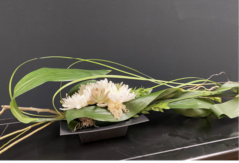 A floral arrangement, featuring a mix of flowers, glossy Aspidistra leaves, arching liriope, and corkscrew willow, is laid out on a counter.