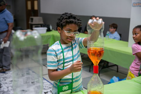 A child in a striped shirt and glasses holds a 2-liter bottle above another 2-liter bottle in an experiment