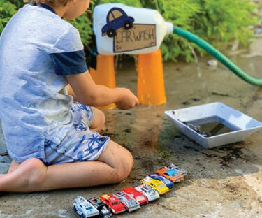A boy kneeling on the wet ground, washing colorful toy cars at his car wash center.