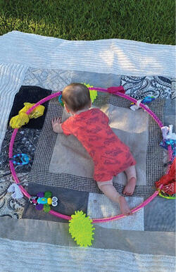 An infant plays in the middle of a pink hula hoop with rattles, bandanas, infant toys, and other soft materials affixed.