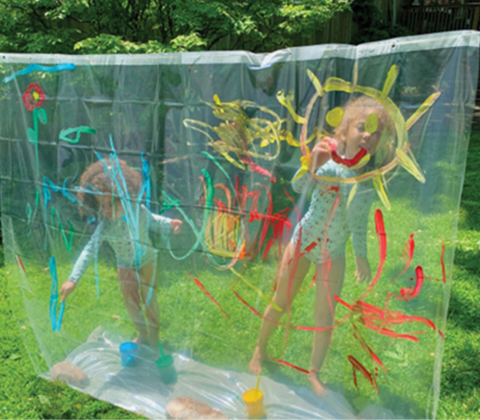 Two young girls paint fun designs on a clear shower curtain that is hanging on a cord outside.