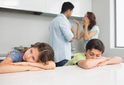 Two kids looking discouraged as two adults are arguing in the background. 