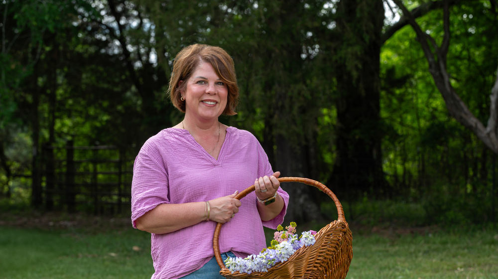 A smiling person standing outside and holding a basket full of colorful flowers.