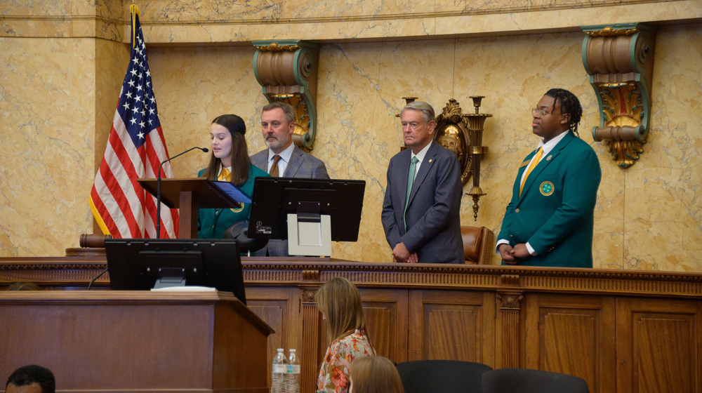 A teenager speaks at a podium as two adults and another teen look on.