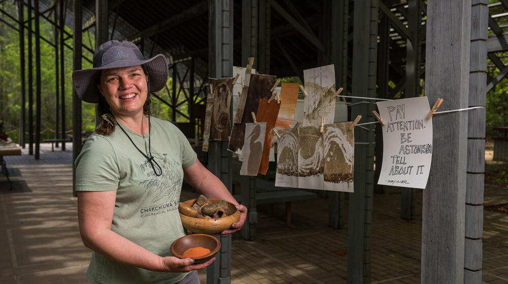 A smiling woman, holding a bowl of rocks in one hand and a bowl of sandy colored dirt in the other, standing in from of paintings hung on a line to dry.