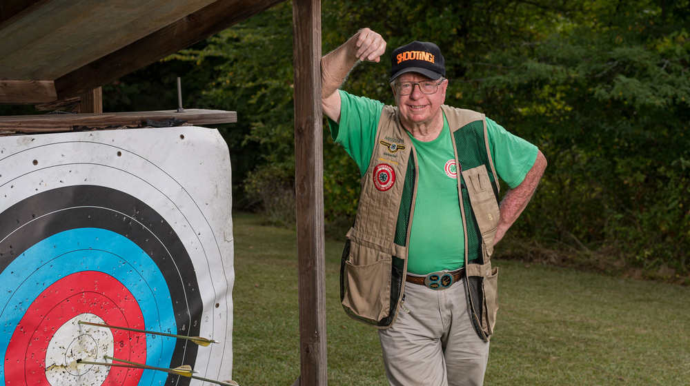 A smiling man wearing a vest and green shirt, standing beside an archery target with three arrows near the bullseye.